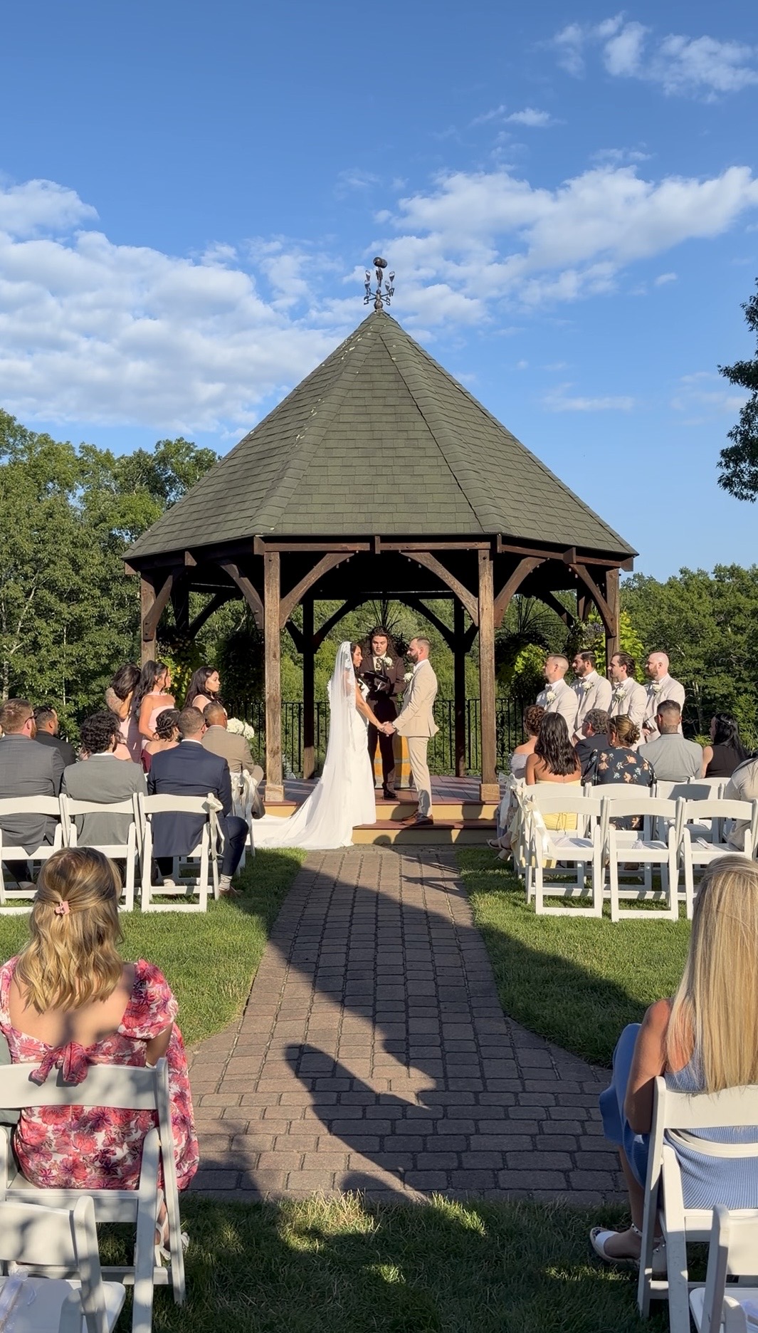 Yazmin & Nick exchanging vows at the gazebo, wedding party and guests seated on either side 
