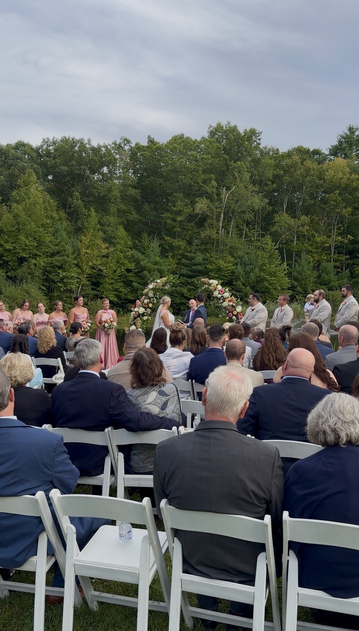 "Outdoor pond ceremony at Zorvino Vineyards with guests seated in white chairs, bridesmaids in pink, and the couple standing beneath twin floral arches"
