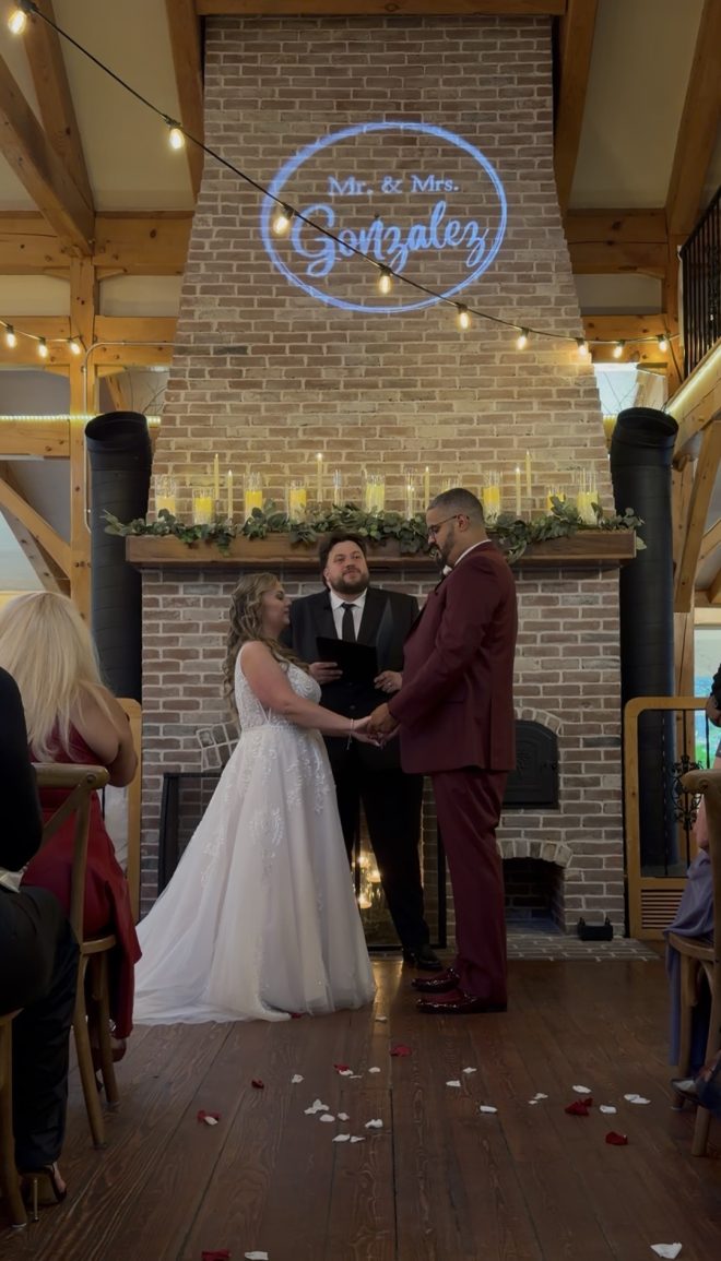 "Fireside fall vineyard wedding ceremony at Zorvino Vineyards: Emily Arsenault and Adrian Gonzalez hold hands before a candlelit brick fireplace beneath a Mr. and Mrs. Gonzalez monogram"