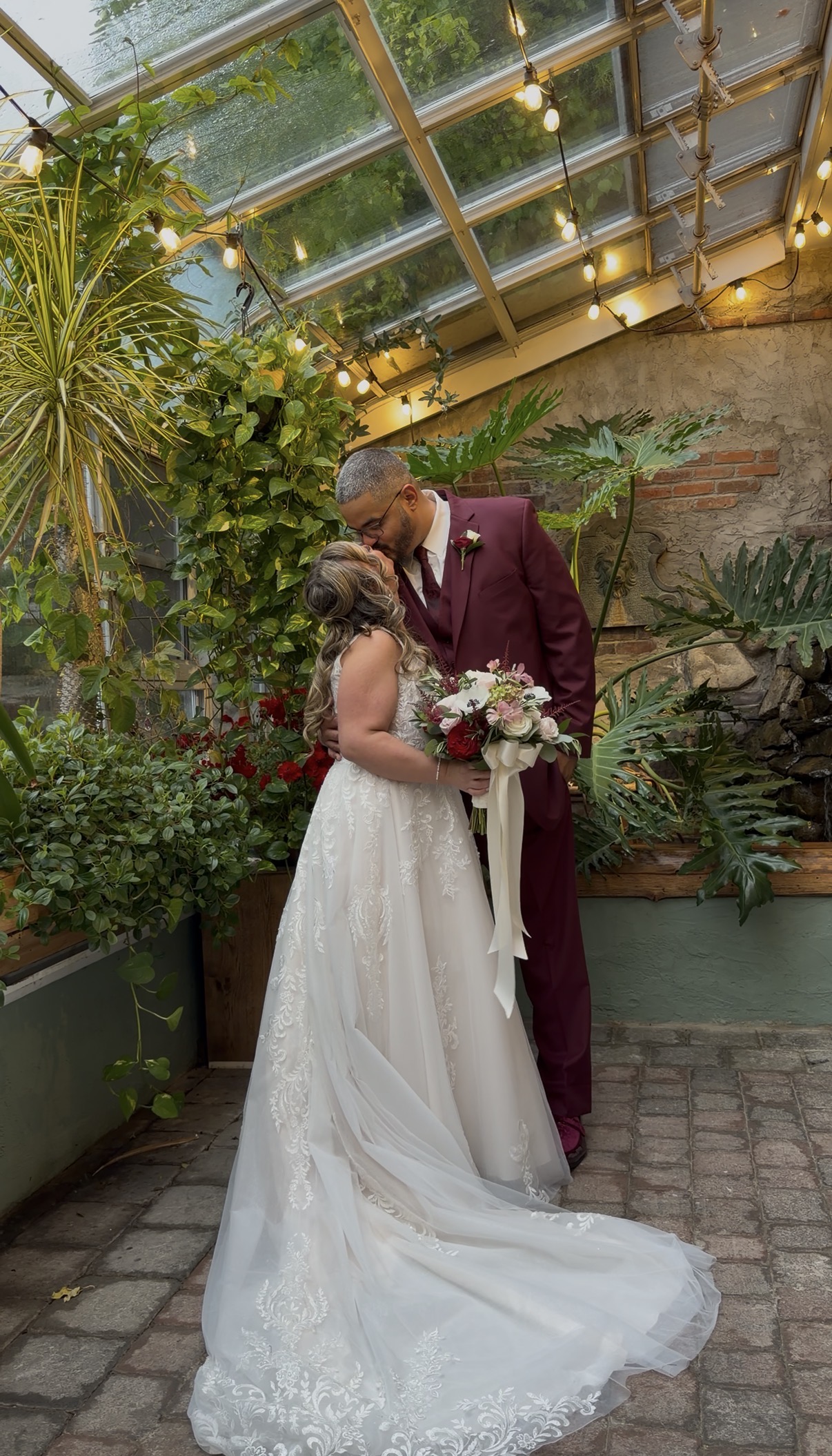 "Emily and Adrian kiss in the Zorvino Vineyards greenhouse conservatory under a glass roof with string lights, hanging plants, and red flowers"