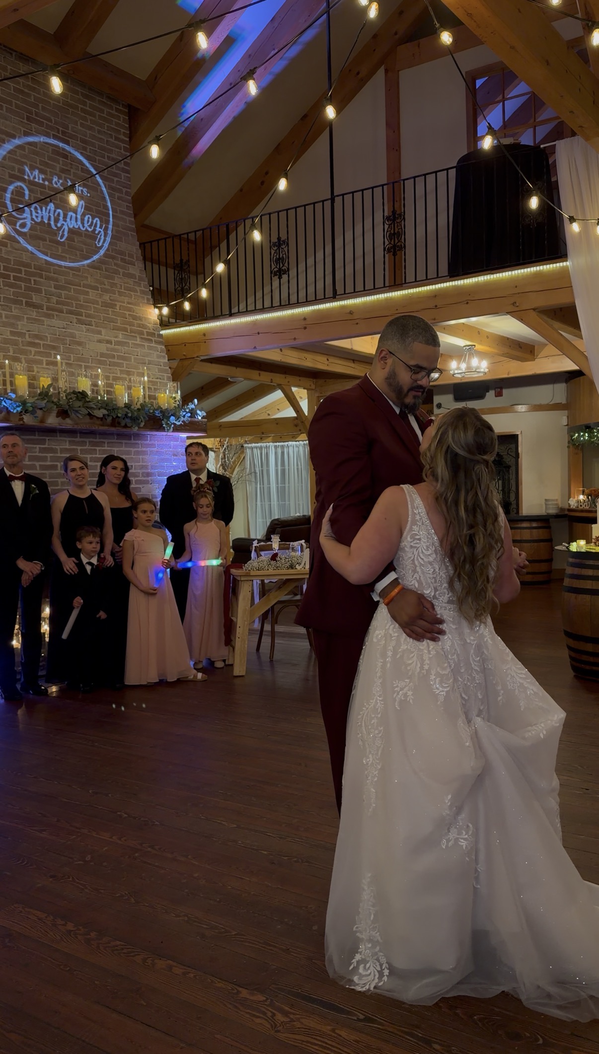 "Emily and Adrian share their first dance in front of the Zorvino fireplace as the wedding party and flower girls with glow sticks watch beneath the projected monogram"