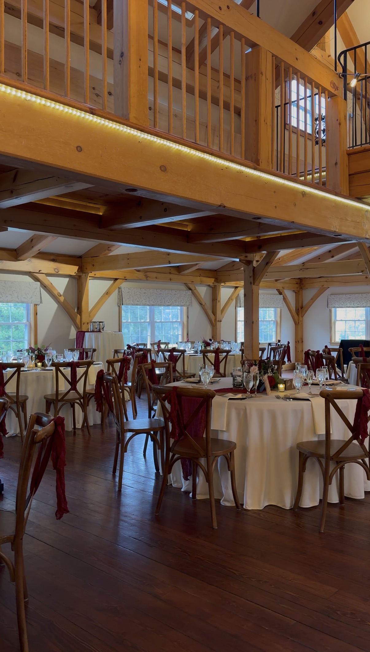 "Reception tables at Zorvino Vineyards with ivory linens, maroon chair sashes on wooden cross-back chairs, gold chargers, and floral centerpieces beneath a timber-frame loft"