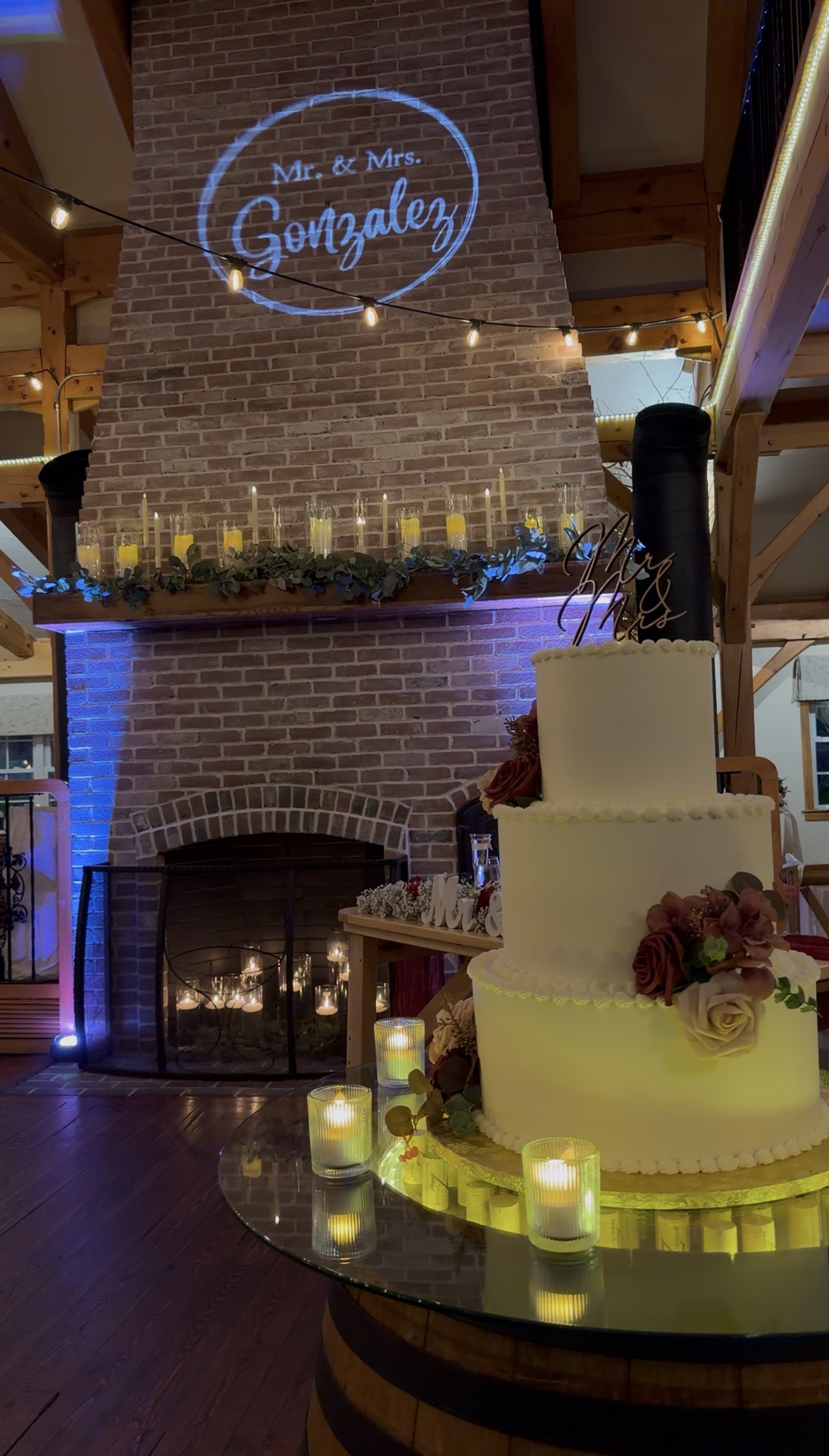 "Three-tier white wedding cake with burgundy roses and a gold Mr and Mrs topper on a candlelit cork barrel display beside the brick fireplace at Zorvino Vineyards"