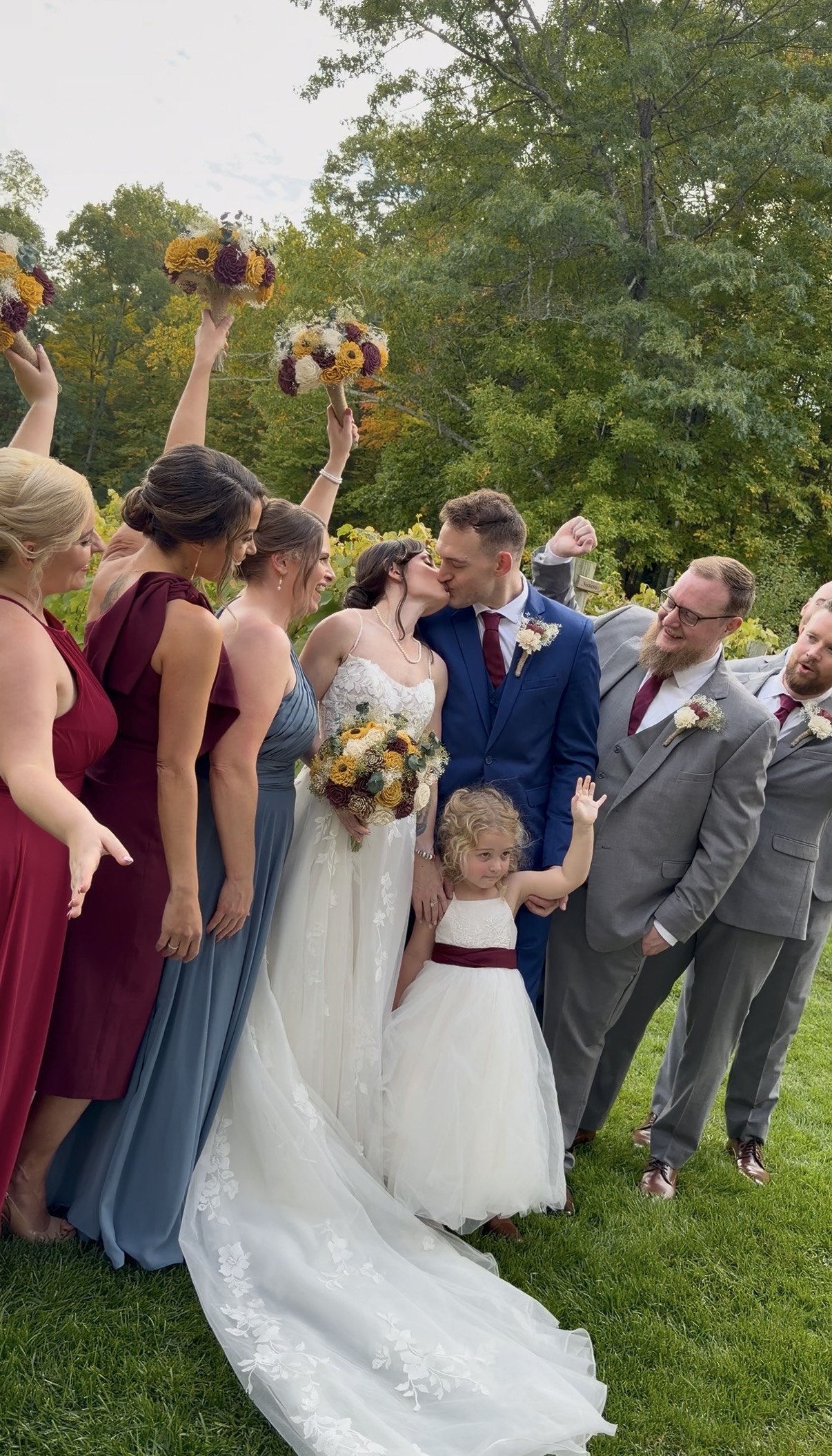 "Bridesmaids in burgundy and slate blue raise sunflower bouquets as Courtney and Andrew kiss among the wedding party and a waving flower girl in the Zorvino vineyard"