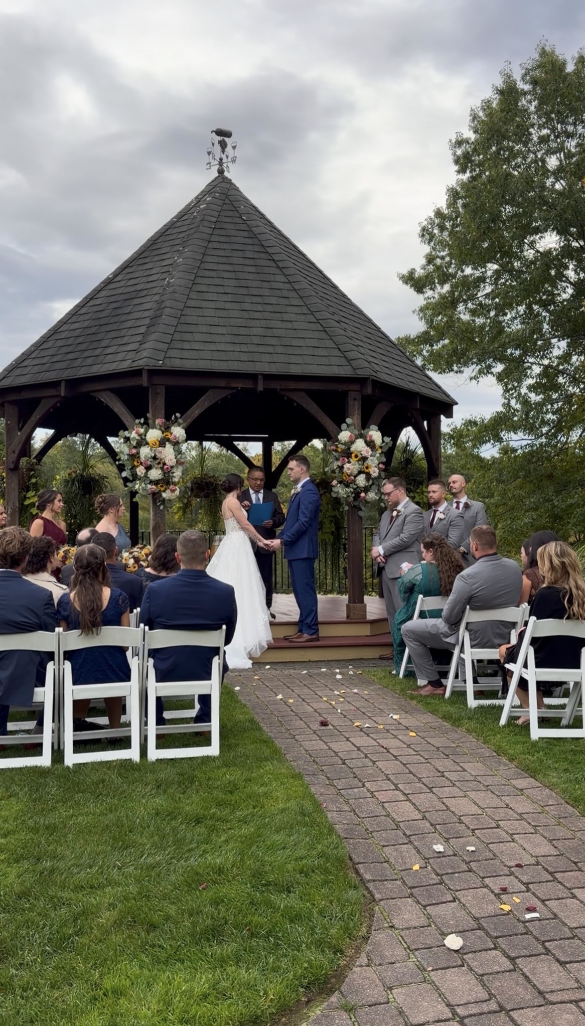 "Courtney and Andrew hold hands at the Zorvino Vineyards gazebo during their outdoor ceremony as guests watch from white chairs along a brick aisle scattered with petals"