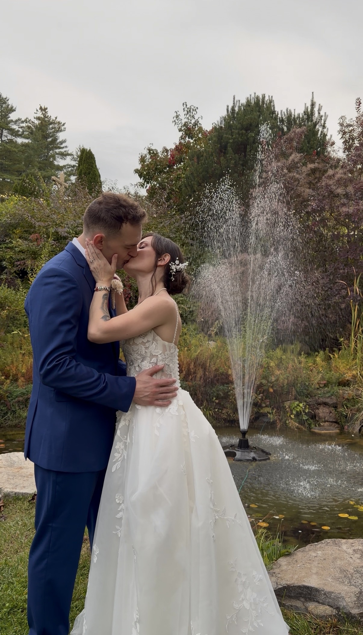 "Courtney and Andrew kiss beside the koi pond fountain at Zorvino Vineyards with autumn shrubs and evergreens behind them"