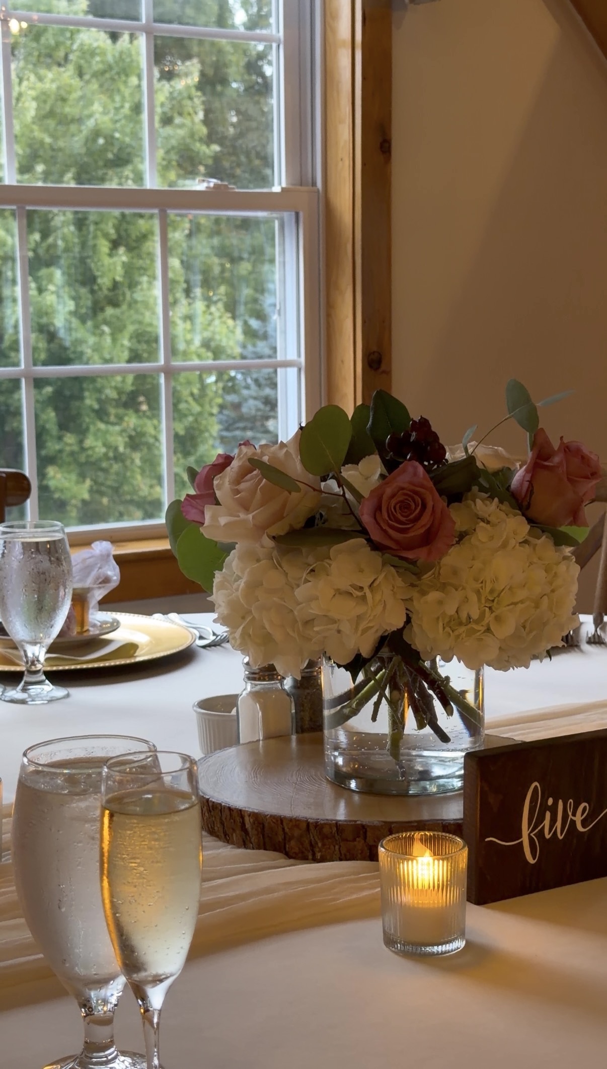 "Centerpiece of white hydrangea, mauve roses, eucalyptus, and berries in a glass vase on a wood slab beside a script table number and votive candle at Zorvino Vineyards"