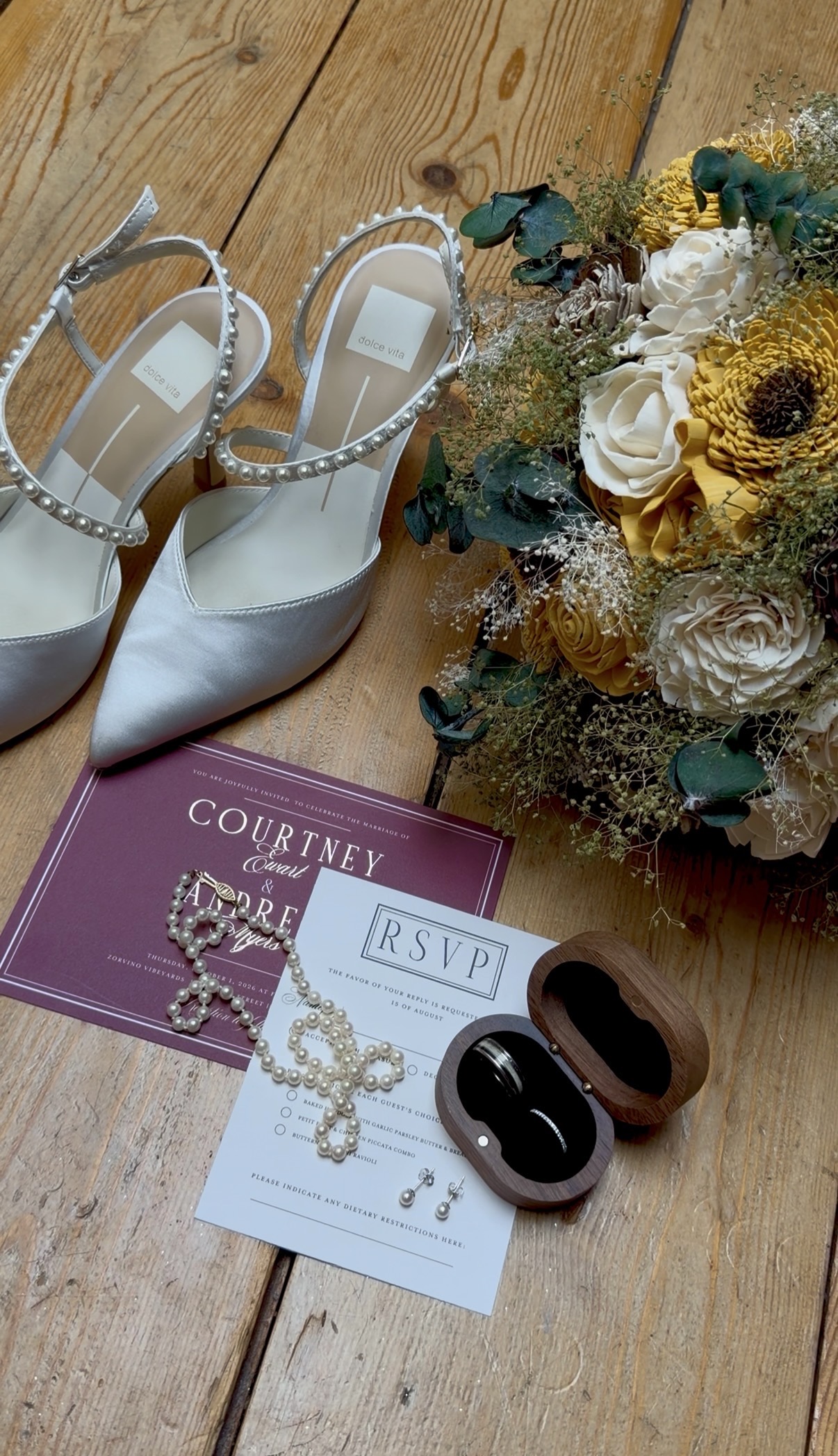 "Burgundy wedding invitation for Courtney Ewart and Andrew Myers with RSVP card, pearl necklace, pearl earrings, pearl-strap bridal heels, wooden ring box, and sunflower bouquet on a wood floor"