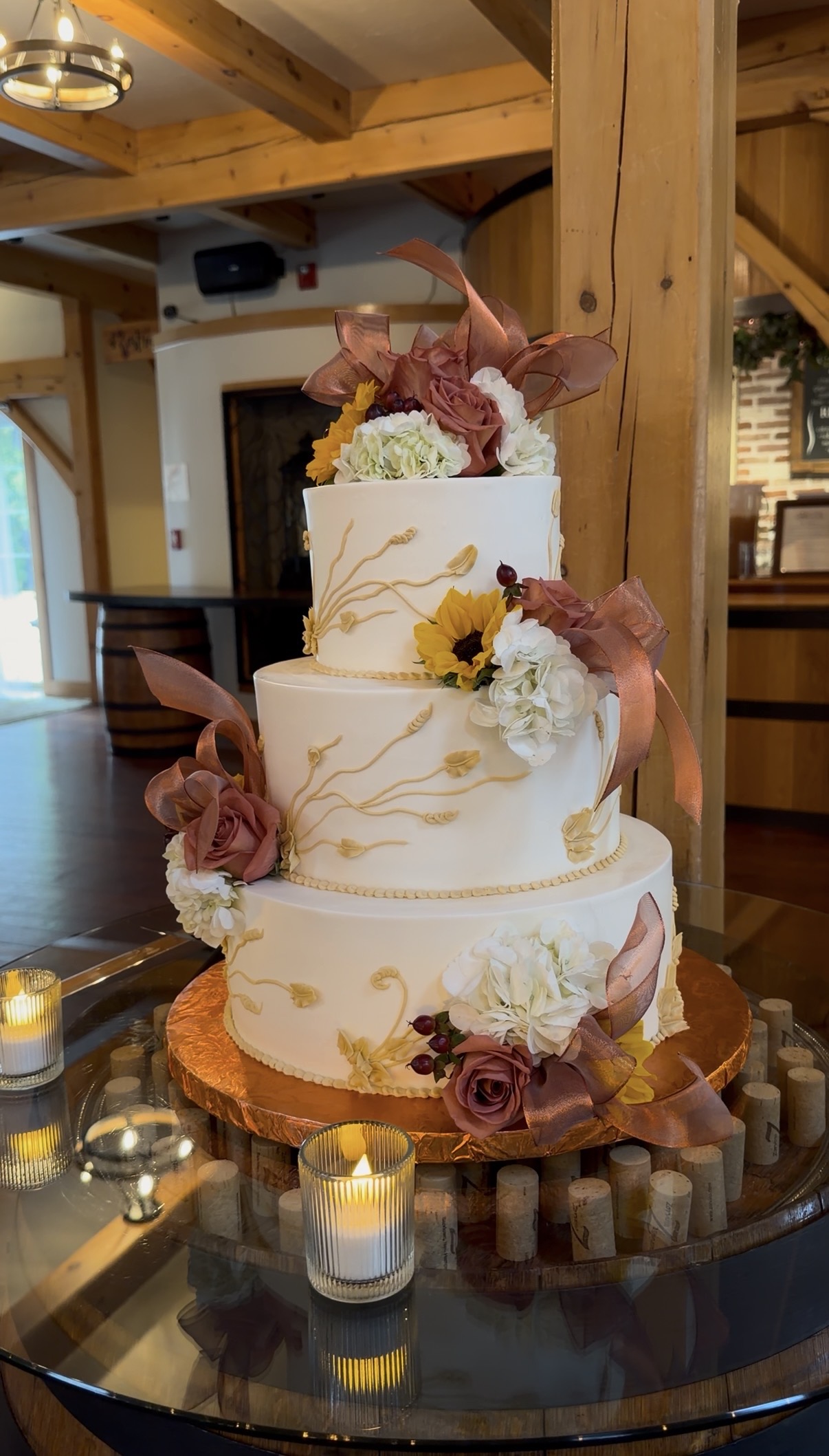 "Three-tier white wedding cake with gold piped wheat, sunflowers, dusty rose roses, hydrangea, and copper ribbon on a cork-filled barrel table at Zorvino Vineyards"