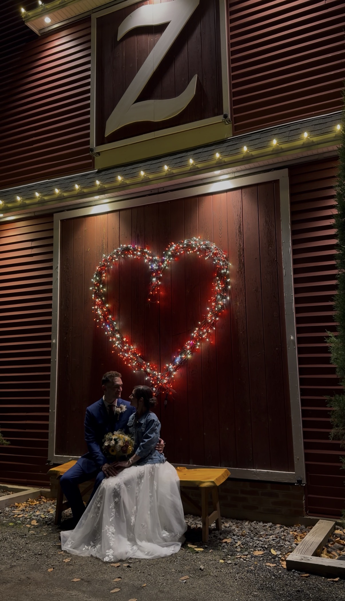 "Courtney in a denim jacket over her wedding gown sits with Andrew on a bench beneath a lit heart on the red barn door at Zorvino Vineyards at night"