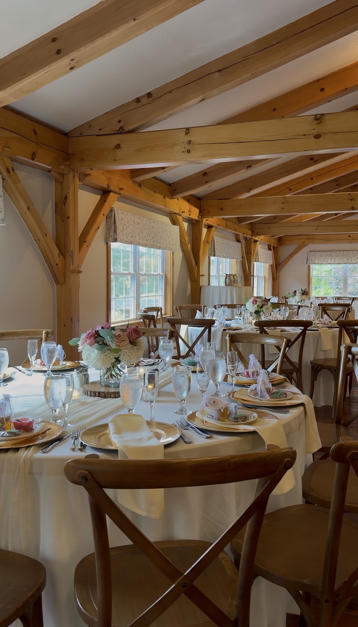 "Reception tables at Zorvino Vineyards with ivory linens, gold beaded chargers, hydrangea and rose centerpieces on wood slabs, and wooden cross-back chairs beneath exposed timber beams"