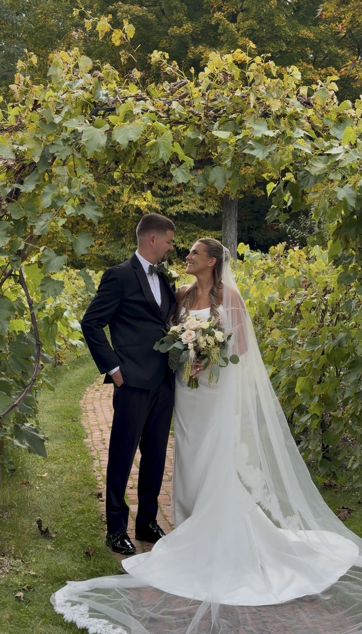 "Olive green wedding at Zorvino Vineyards: Sophia Holman and Scott Donahue smile at each other on a brick path beneath a grapevine arbor in October"