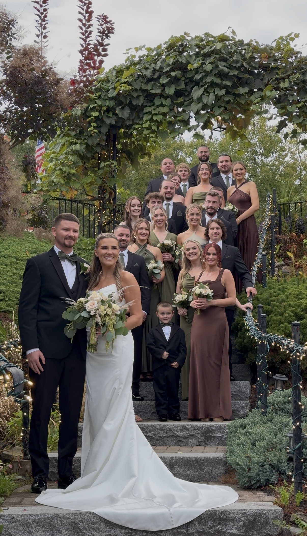 Bride and groom stand at the foot of stone steps under a grapevine arbor with string lights as bridesmaids in olive green and espresso brown satin and groomsmen in black fill the steps behind them.