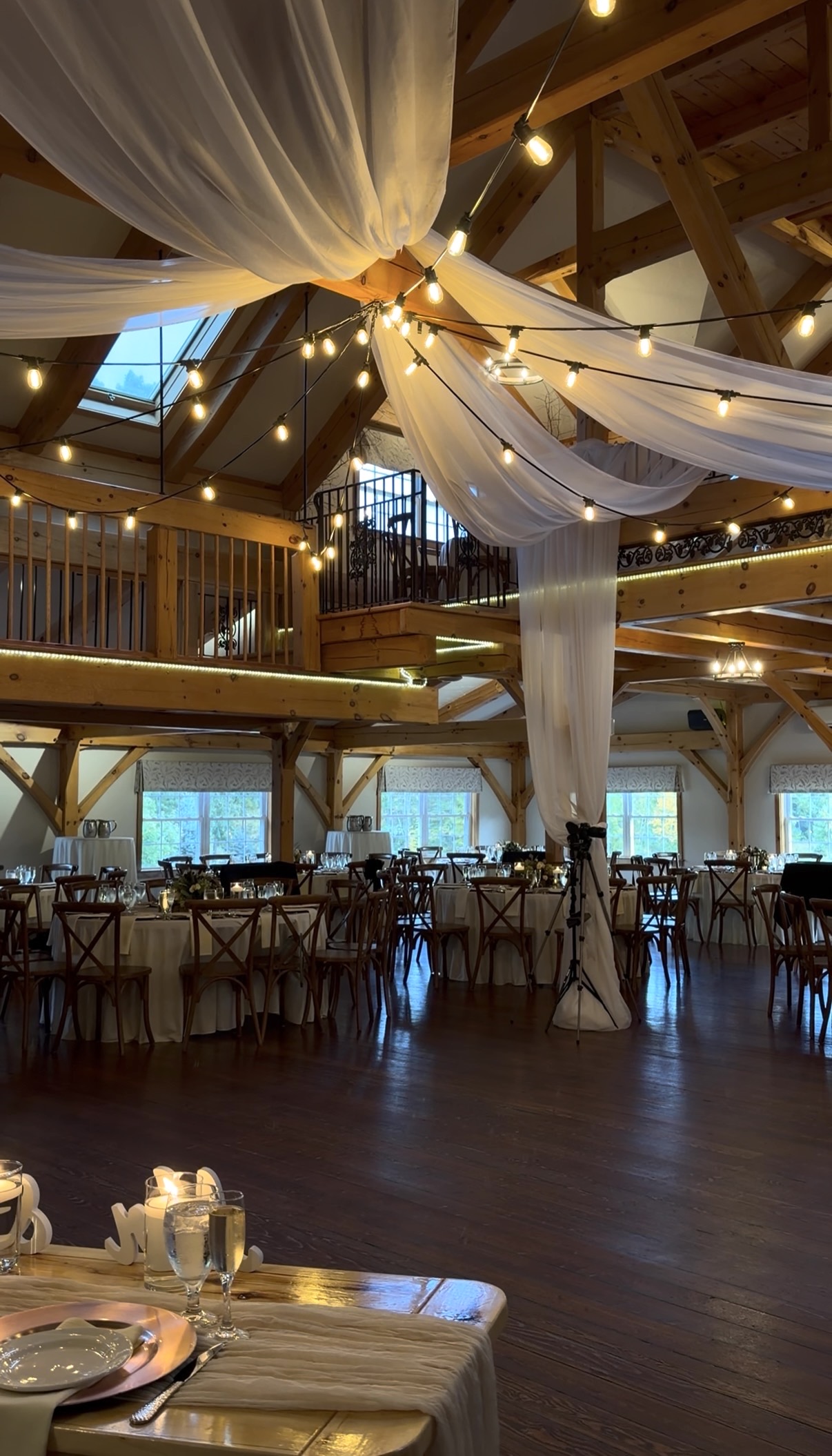 Zorvino Vineyards timber-frame reception hall with sheer ivory ceiling drapery, string lights, a wooden loft, round tables with ivory linens, and a sweetheart table with rose gold chargers in the foreground.