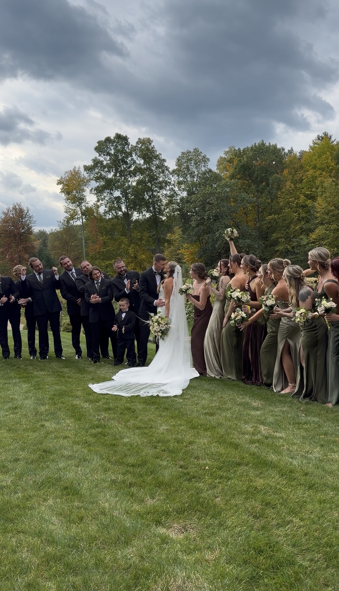 Sophia and Scott kiss on the Zorvino lawn as bridesmaids in olive and espresso satin and groomsmen in black cheer on either side.