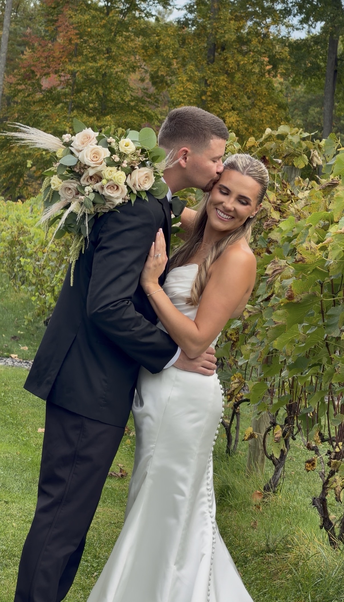 Scott kisses Sophia on the cheek beside the vine rows at Zorvino Vineyards as she holds a bouquet of quicksand roses, pampas grass, and green amaranthus over his shoulder.