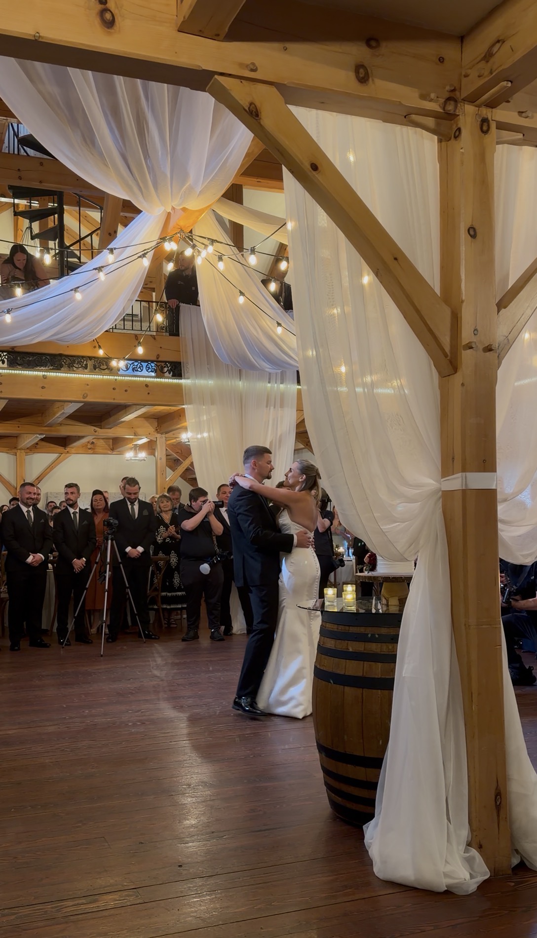 Sophia and Scott share their first dance beneath ivory drapery and bulb lights at Zorvino Vineyards as groomsmen and guests watch from the floor and the loft above.