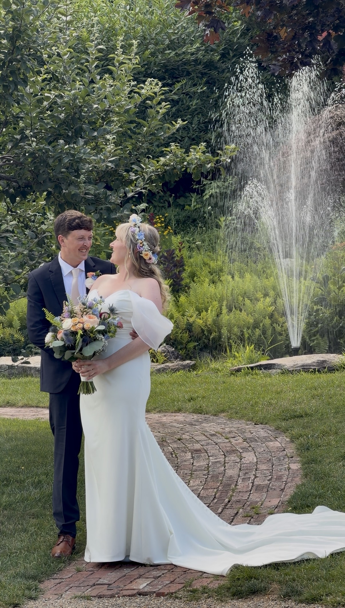 A bride and groom stand together and laugh in front of a fountain.