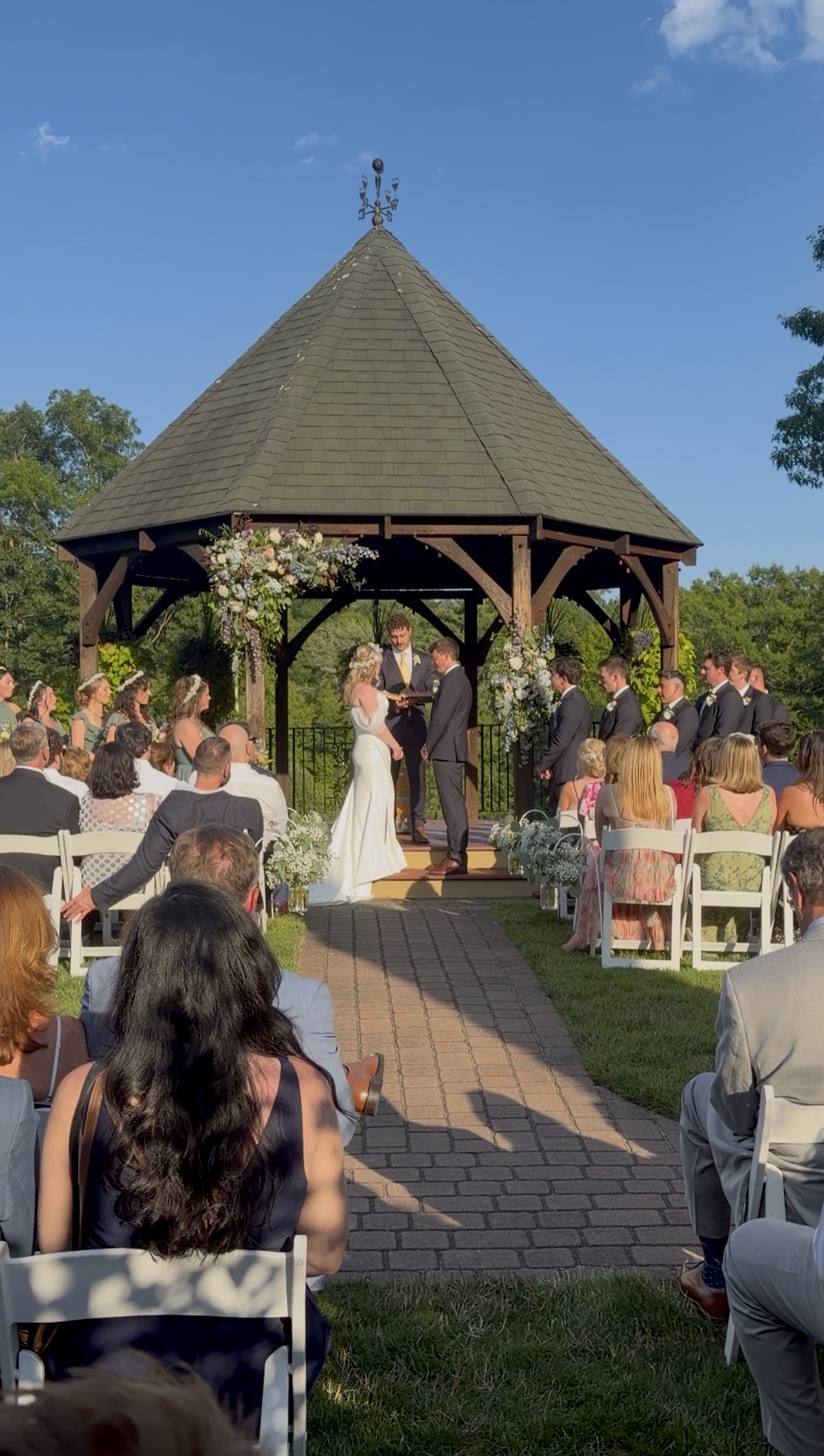 A bride and groom in front of the altar in front of the gazebo with wedding guests watching the ceremony.