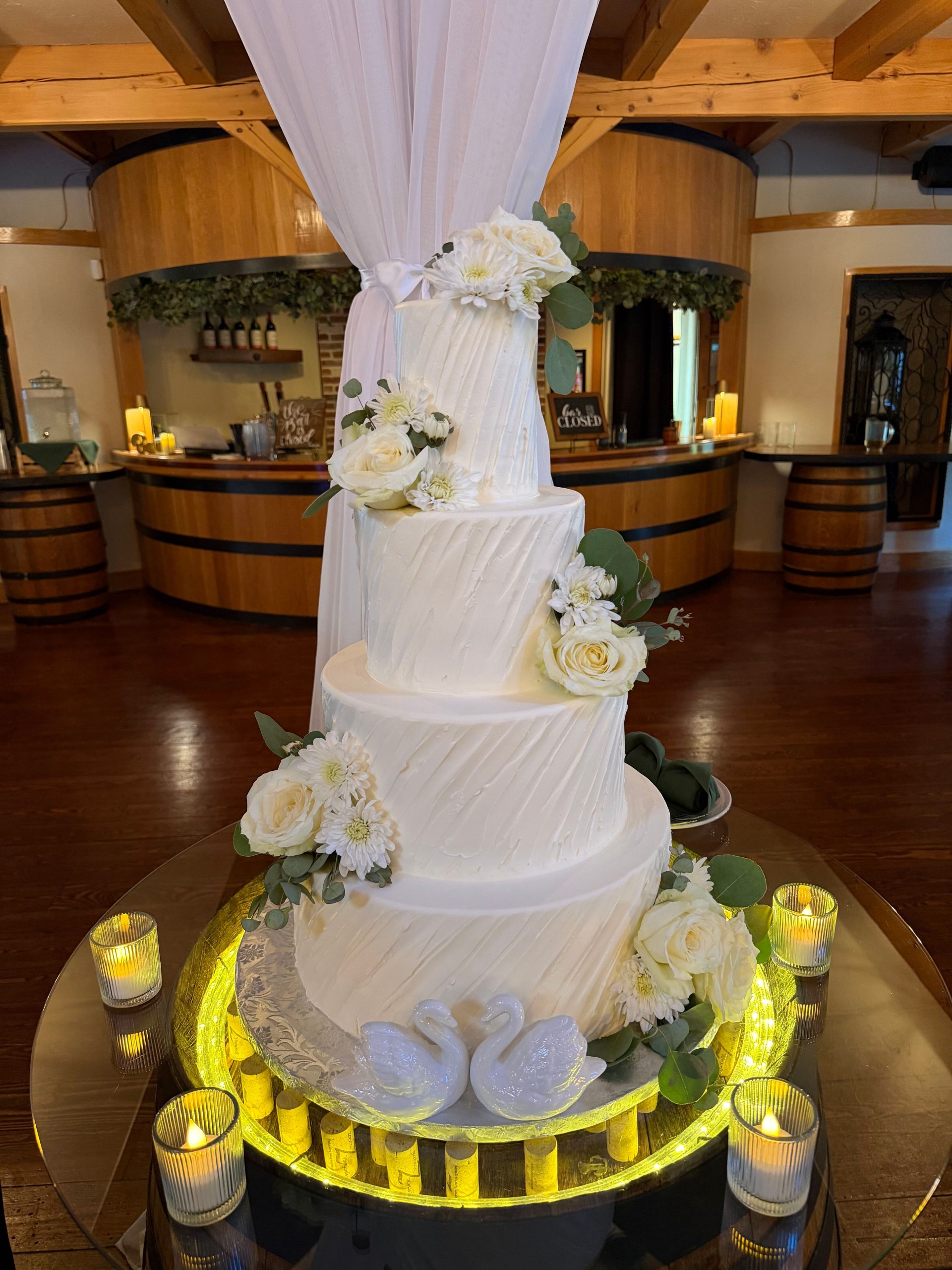 A large 4-tiered wedding cake with white flowers and ceramic swans in the candlelight.