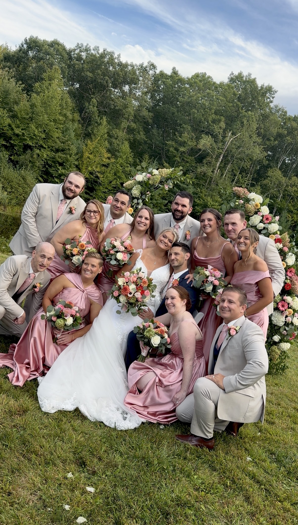 "Wedding party of bridesmaids in dusty rose satin with coral and blush bouquets gathered around the newlyweds on the lawn at Zorvino Vineyards"

