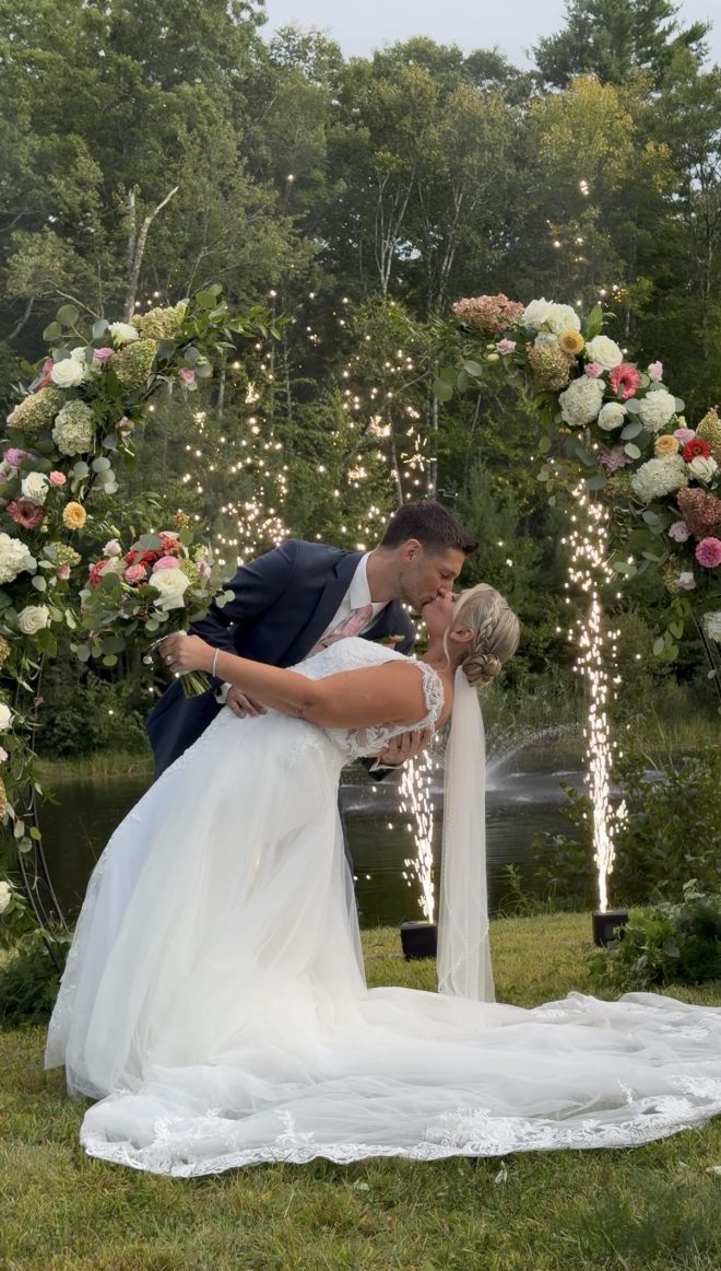 "Travel themed vineyard wedding at Zorvino Vineyards: Khyrstyne Quigley and Michael Pieroni kiss under floral arches at the pond as cold sparks rise behind them"