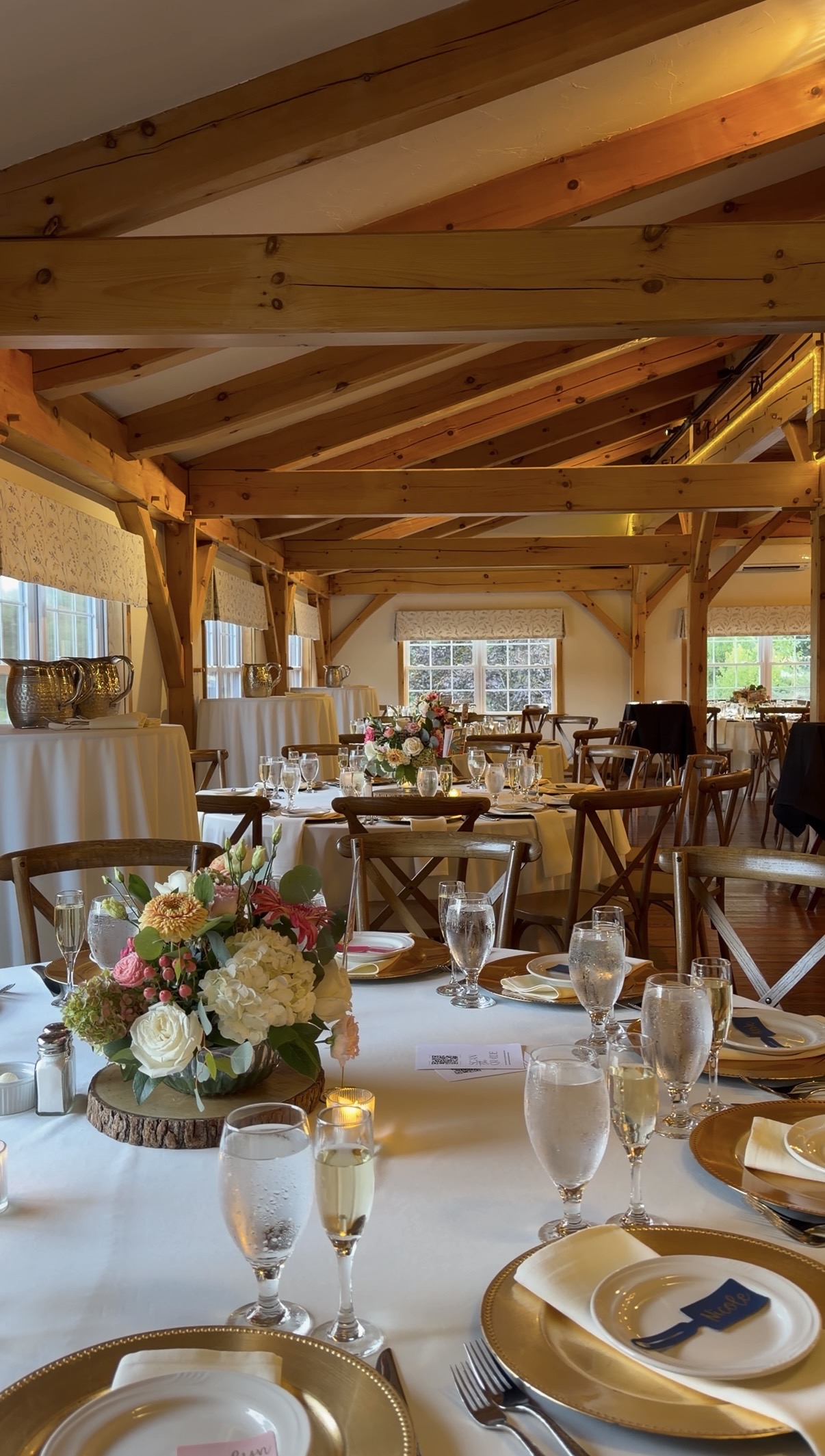 "Reception tables in the post-and-beam barn at Zorvino Vineyards with gold chargers, wood slab centerpieces of hydrangea and roses, and votive candles"
