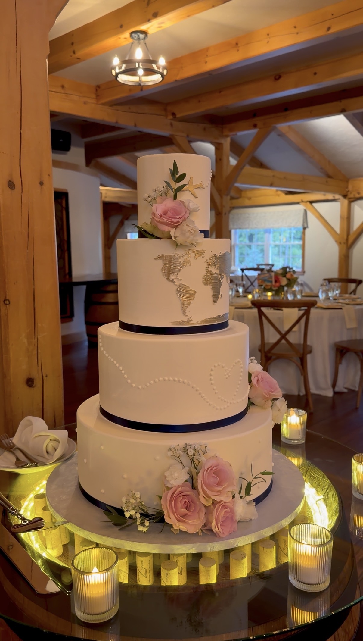 "Four-tier white travel-themed wedding cake with gold world map, piped dotted flight path ending in a heart, gold airplane, navy ribbon, and pink roses"

