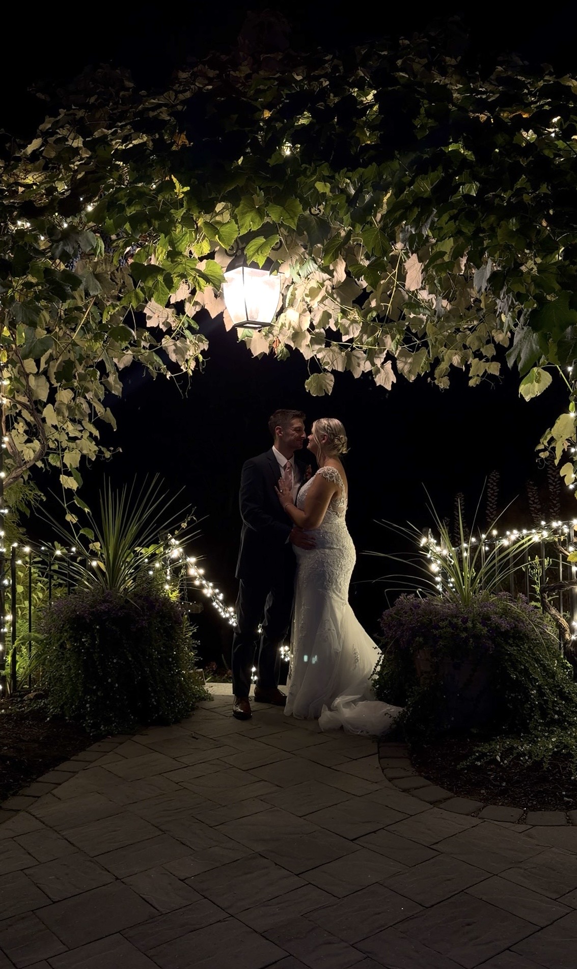 "Khyrstyne and Michael embrace at night beneath a grapevine arbor with a glowing lantern and string lights at Zorvino Vineyards"
