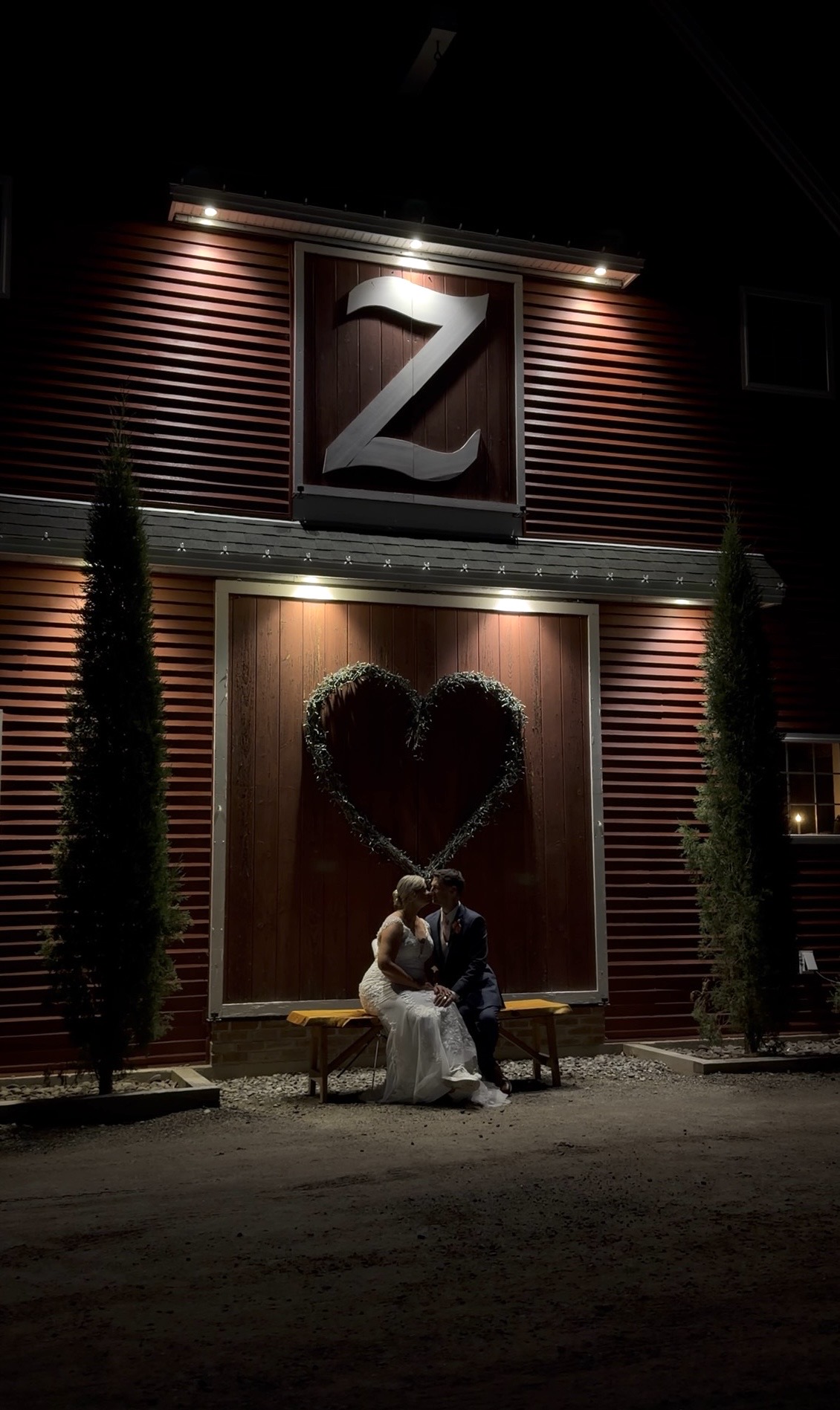 "Newlyweds kiss on a bench at night below the illuminated Z and heart wreath on the red barn at Zorvino Vineyards in Sandown NH"

