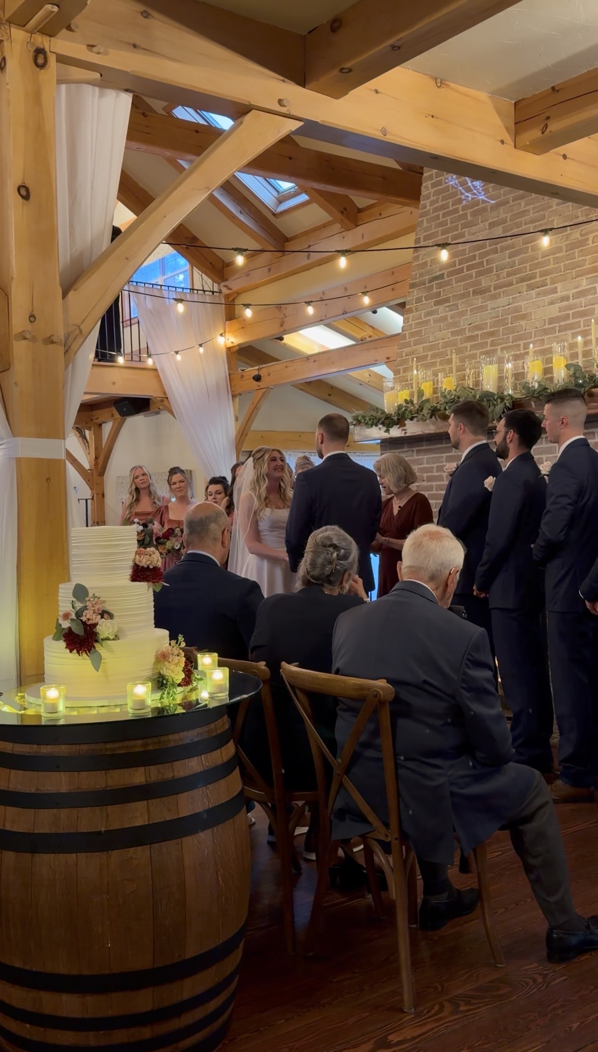 Fireside wedding ceremony at Zorvino Vineyards with the bride facing the groom before a candlelit brick mantel, grandparents seated in front, and a three-tier cake on a wine barrel in the foreground.