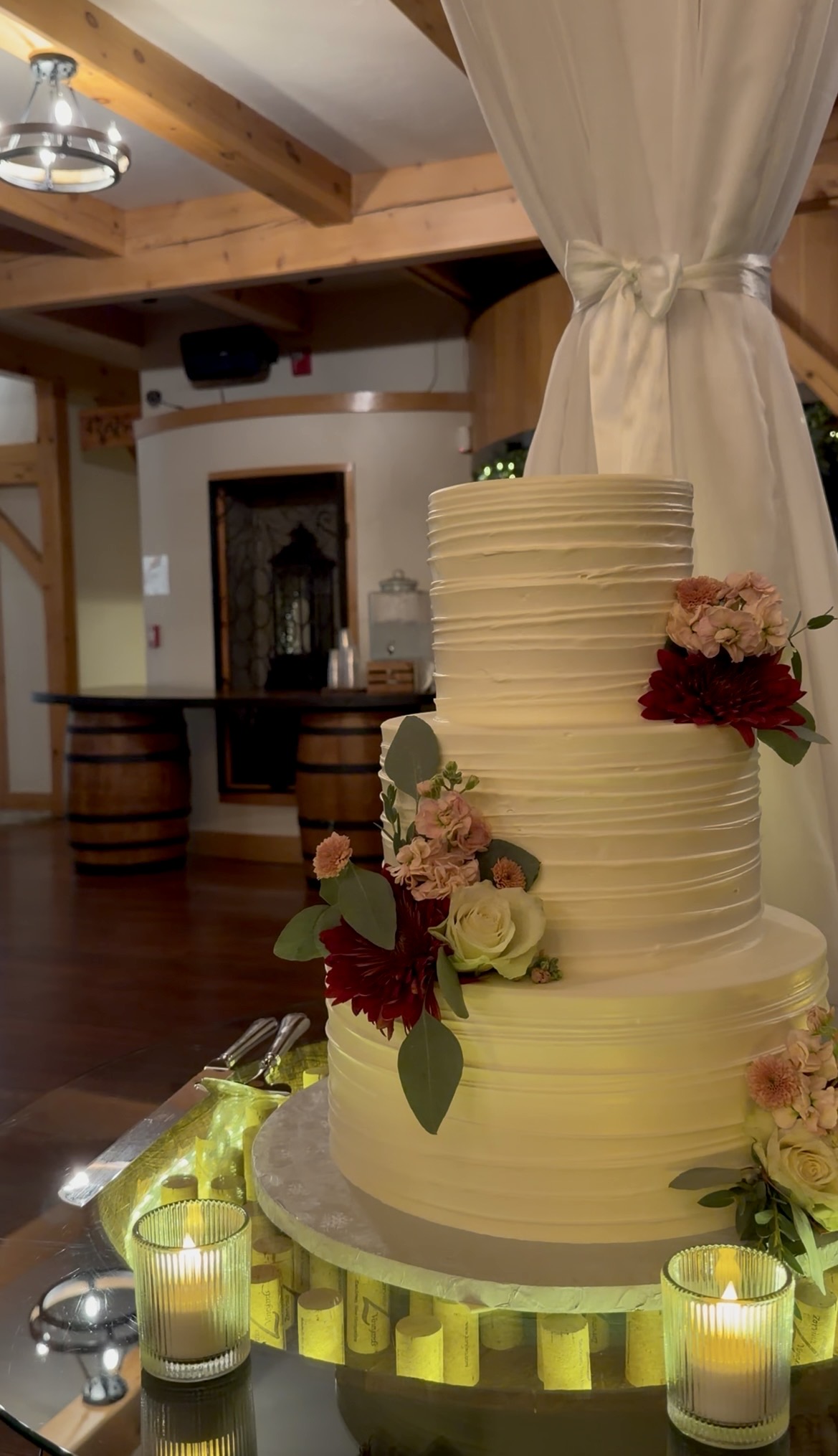 Three-tier textured ivory buttercream wedding cake with burgundy dahlias, blush stock, and ivory roses on a candlelit glass barrel display filled with Zorvino wine corks.