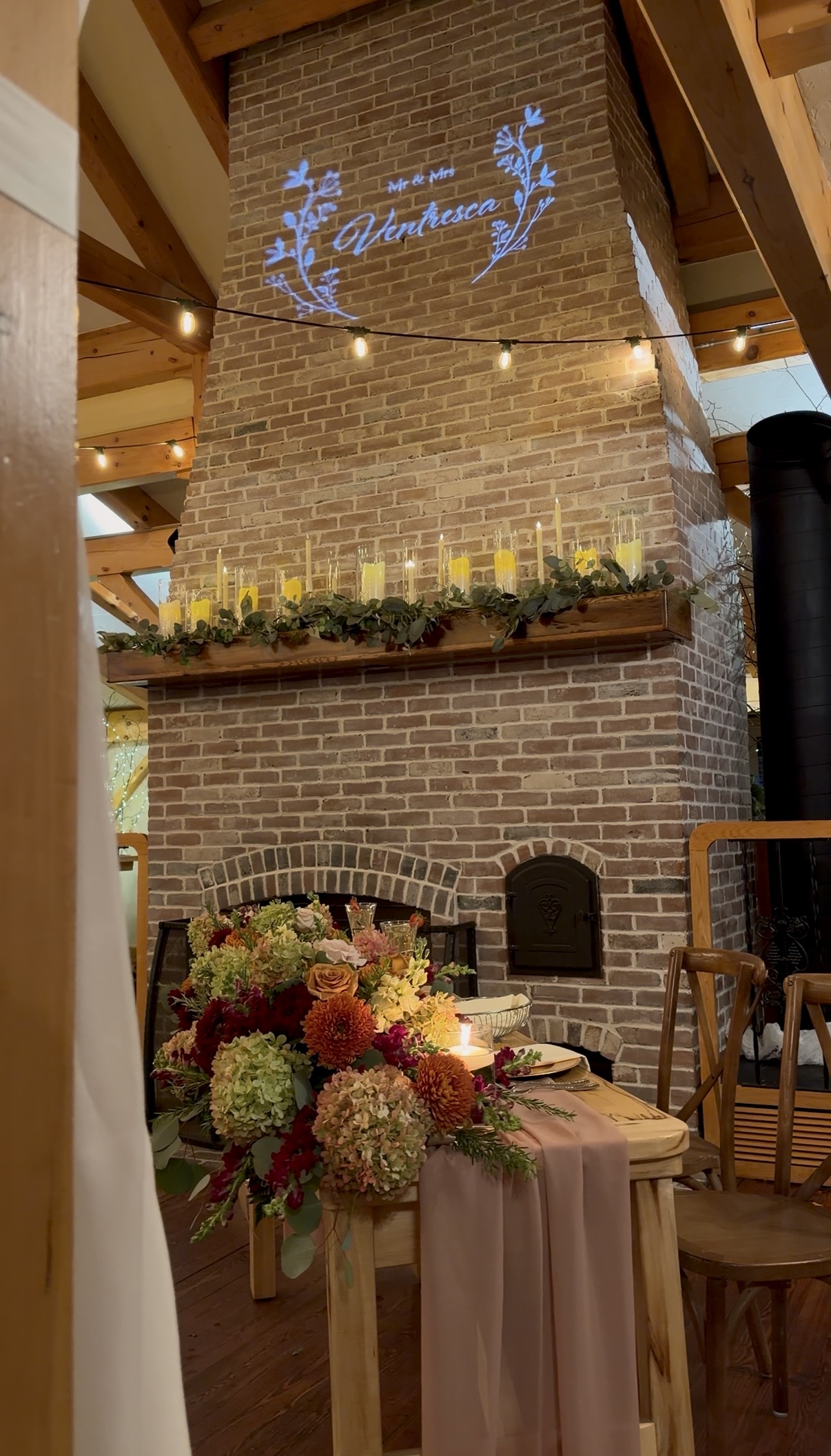 Sweetheart table with a fall floral arrangement and mauve chiffon runner in front of the brick fireplace at Zorvino Vineyards, Mr. and Mrs. Ventresca monogram projected above.