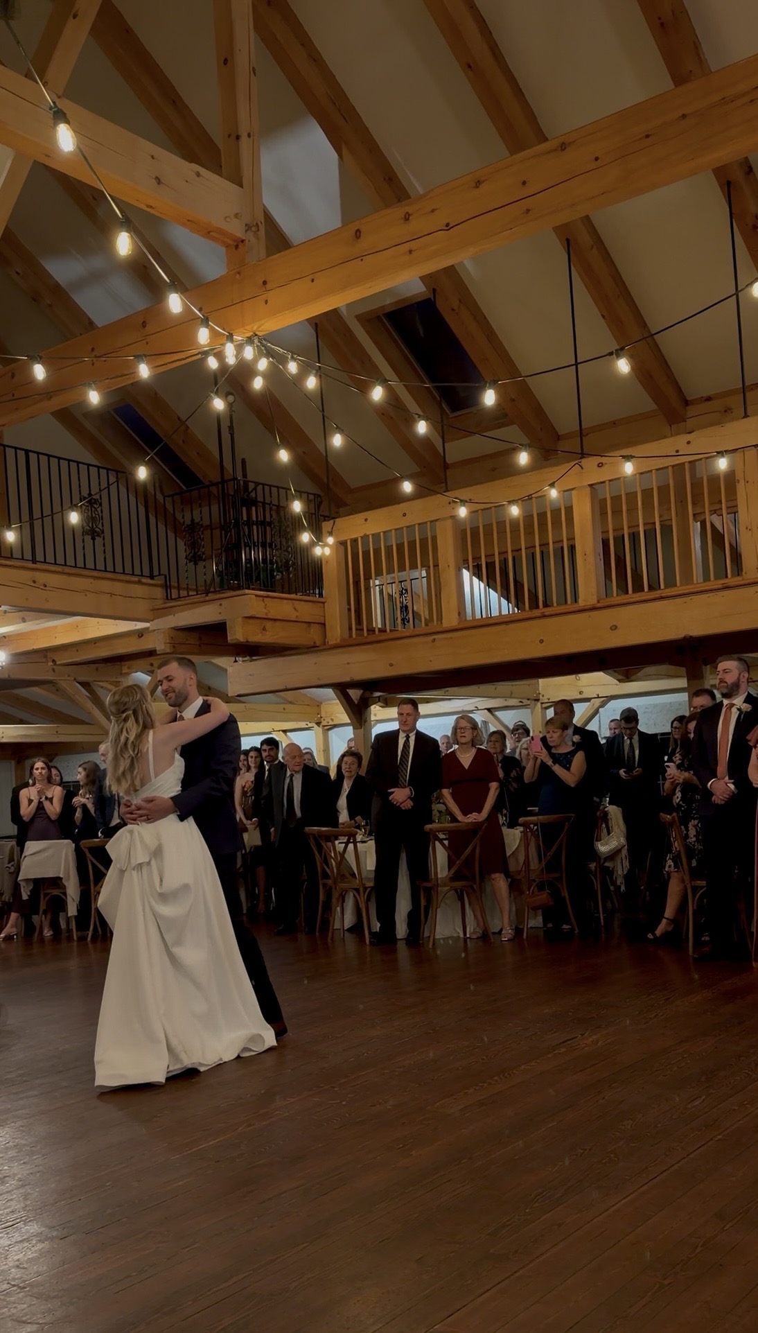 Meghan and Alex share their first dance on the wood floor of the Zorvino Vineyards reception hall under string lights and a timber loft as guests watch.
