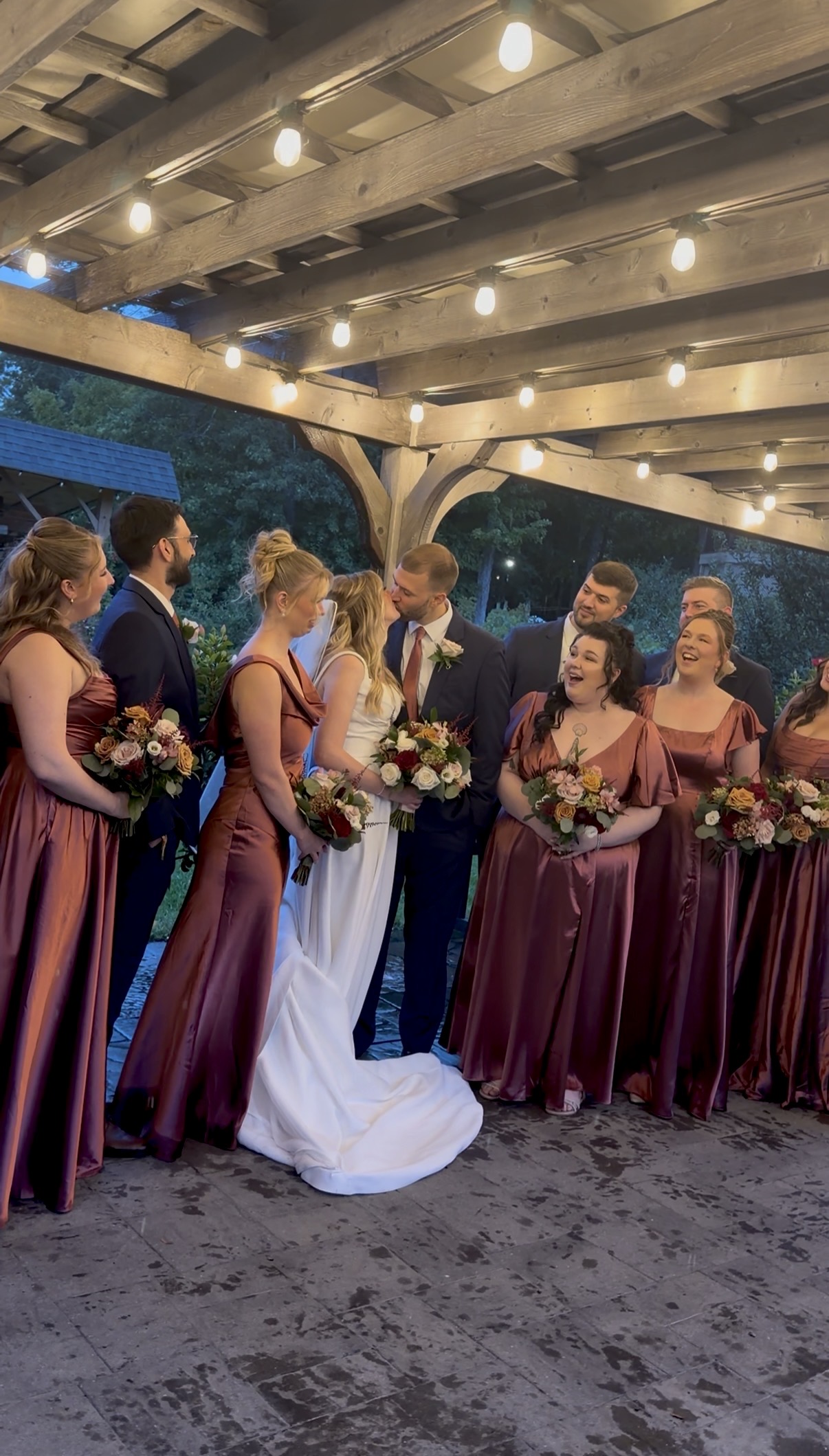 Bridal party in mauve satin gowns and navy suits laughs as the bride and groom kiss under the string-lit pergola on the Zorvino Vineyards patio.