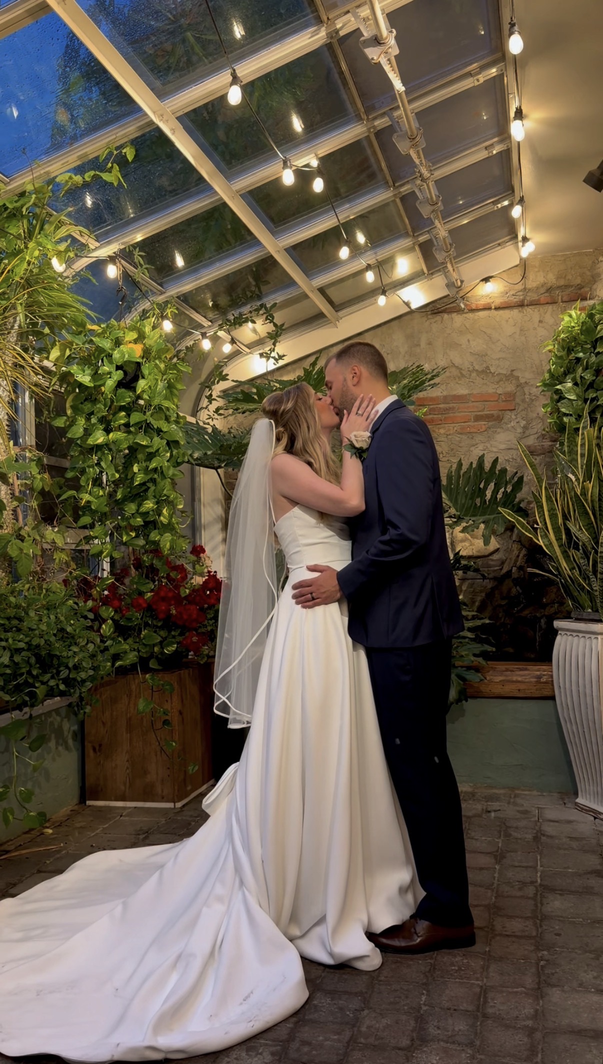 Meghan and Alex kiss in the Zorvino Vineyards greenhouse conservatory beneath a glass roof with string lights, hanging pothos, and red geraniums.