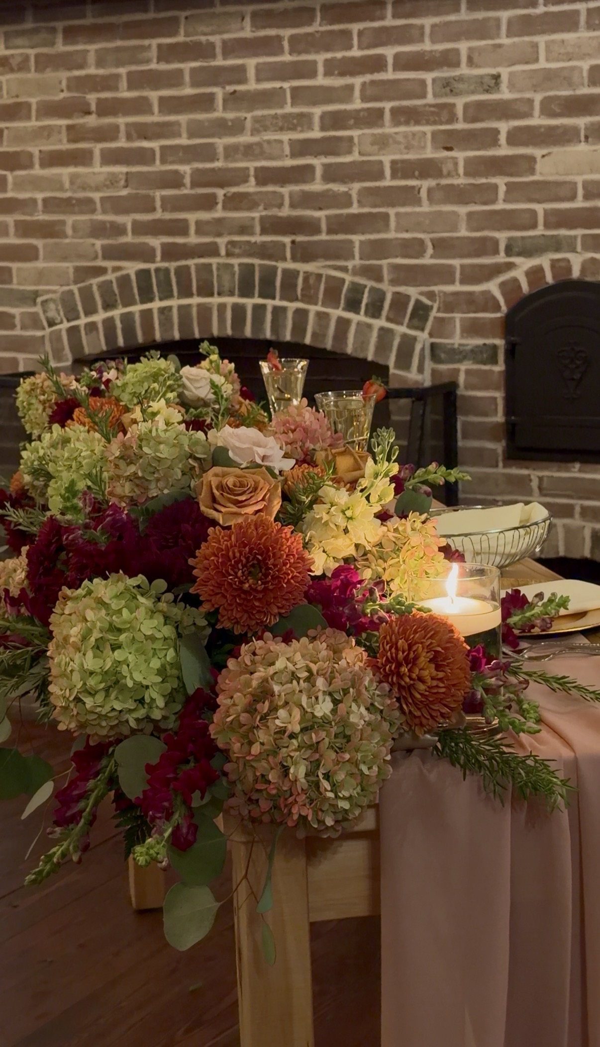 Close view of a sweetheart table arrangement of green and pink hydrangea, rust chrysanthemums, burgundy dahlias, toffee roses, and a floating candle against a brick hearth.