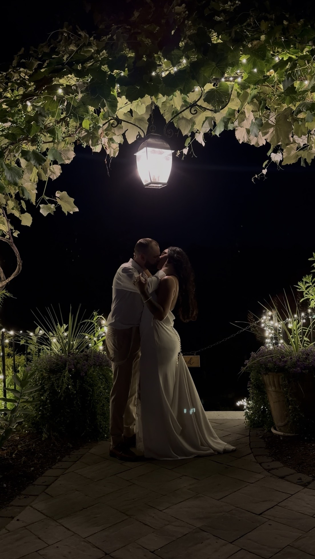 Yazmin & Nick kissing under a lantern beneath the vine-draped patio archway at night