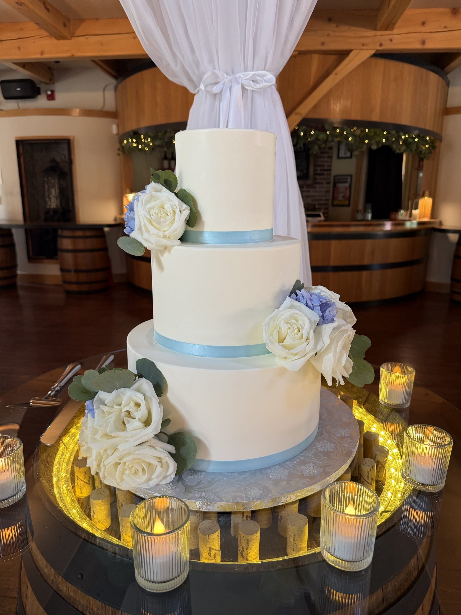 Three-tier white wedding cake with pale blue ribbon and roses at a family vineyard wedding at Zorvino Vineyards A three-tier cake in ivory and pale blue, glowing atop a ring of wine corks.