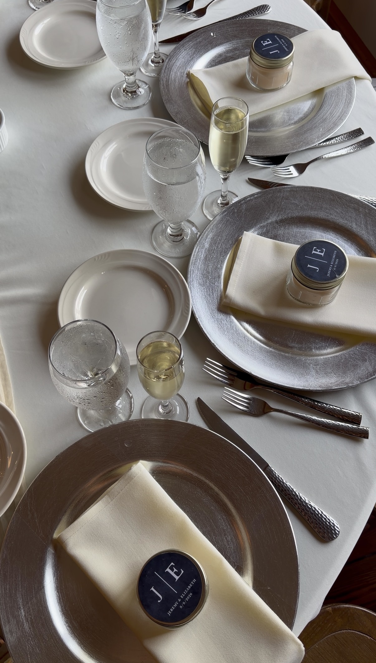 Silver charger place settings with personalized candle favors at a family vineyard wedding at Zorvino Vineyards Silver chargers, ivory linens, and a little candle to take home.