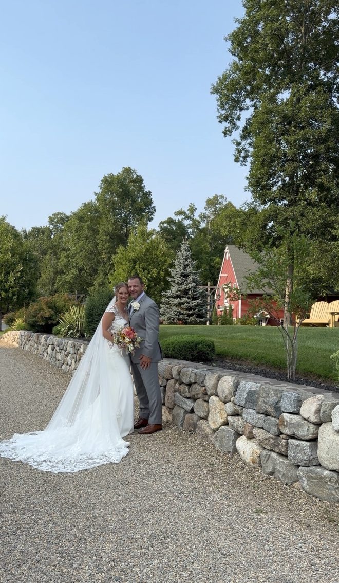 Elizabeth and Jeremy by the stone wall with Zorvino's red barn behind them at their family vineyard wedding in Sandown, NH